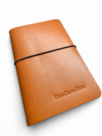 Caderno artesanal em couro genuíno castanho, capa reutilizável com elástico e gravação personalizada, fotografado em ambiente minimalista com luz natural.
